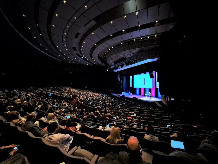 People sitting in a packed auditorium with a presentation taking place on stage
