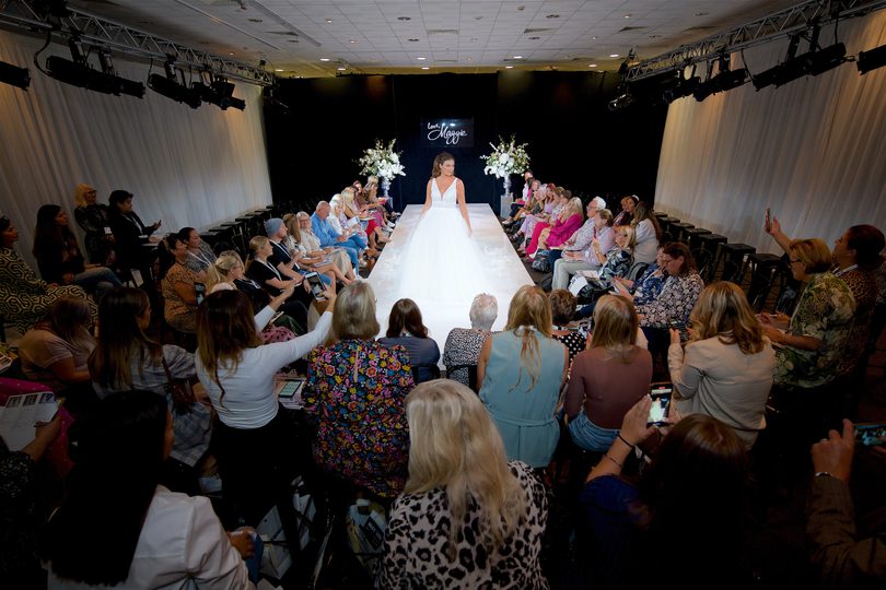 A model wearing a white wedding dress posing on a runway surrounded by an audience taking photos with their phones