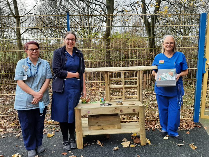 Three hospital staff standing next to a wooden toy kitchen in an outdoor setting.