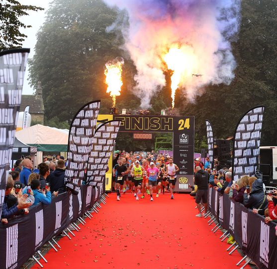 A group of runners dashes on a red carpet, with flames shooting up from above, and spectators cheering from both sides of the route.